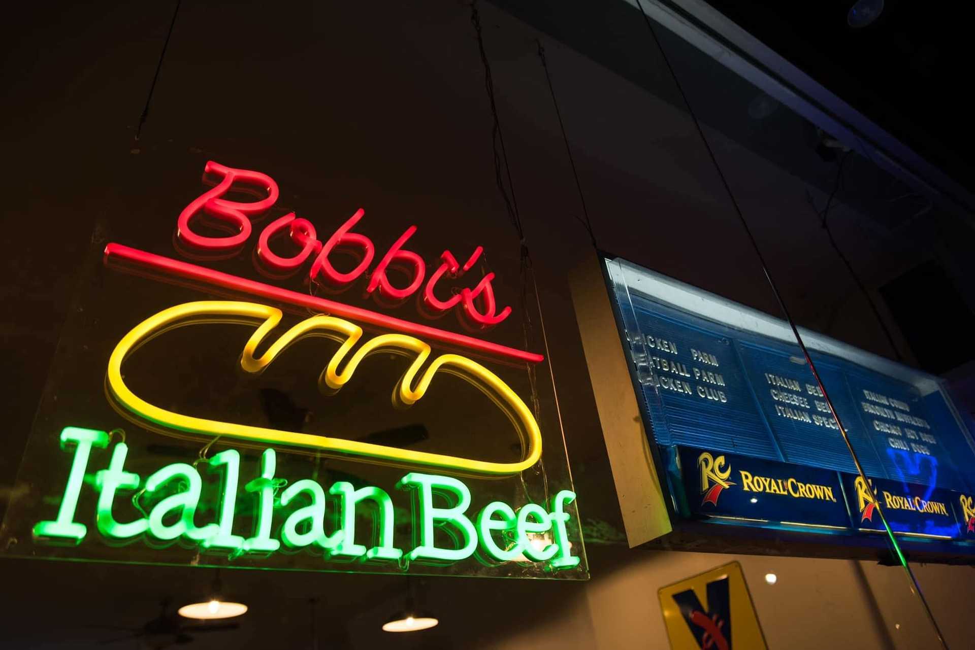 Neon sign reads "Bobby's Italian Beef" above a sandwich icon with menu boards in the background.